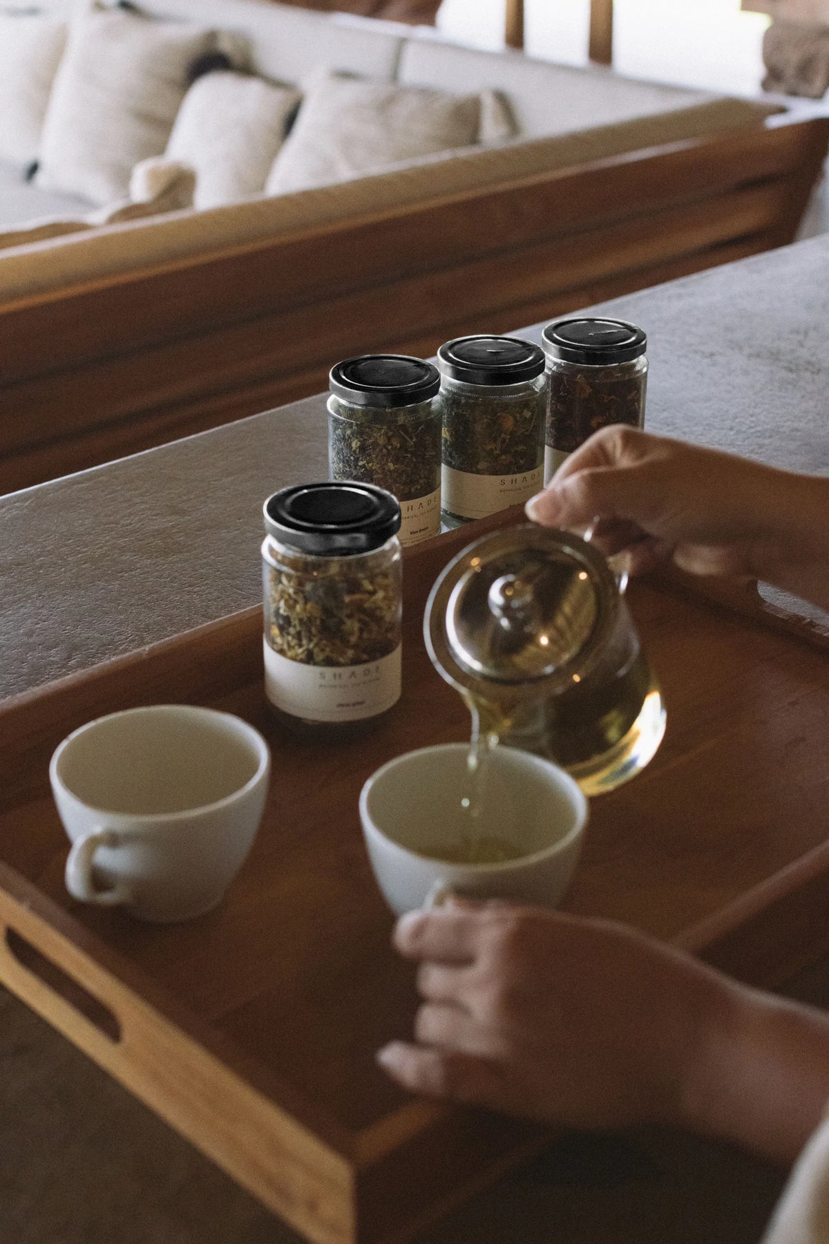 Person pouring Shade Botanicals Tea Blend from a teapot into a cup on a wooden tray with jars of tea leaves