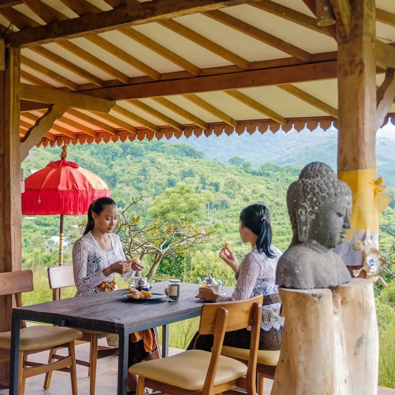 Two women sitting at a table with a scenic view, a red umbrella, and a statue