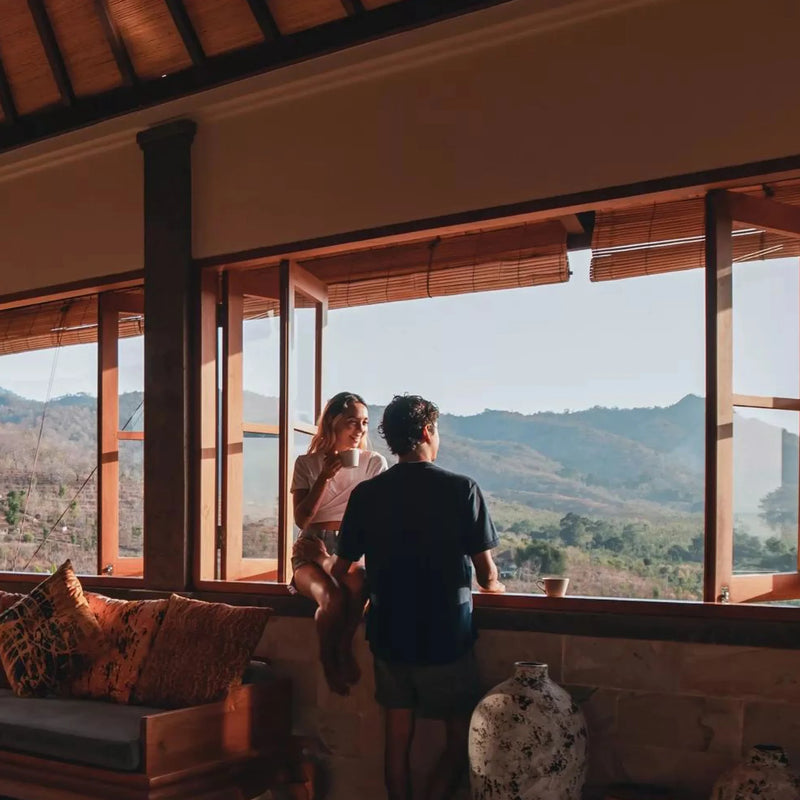 Two people sitting on a couch inside a house with large windows overlooking a scenic view of mountains