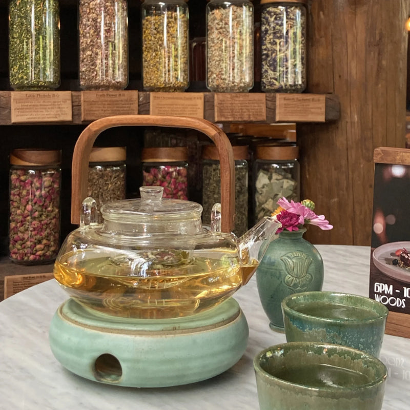 Tea set with a glass teapot on a green warmer, surrounded by jars of tea leaves in the background