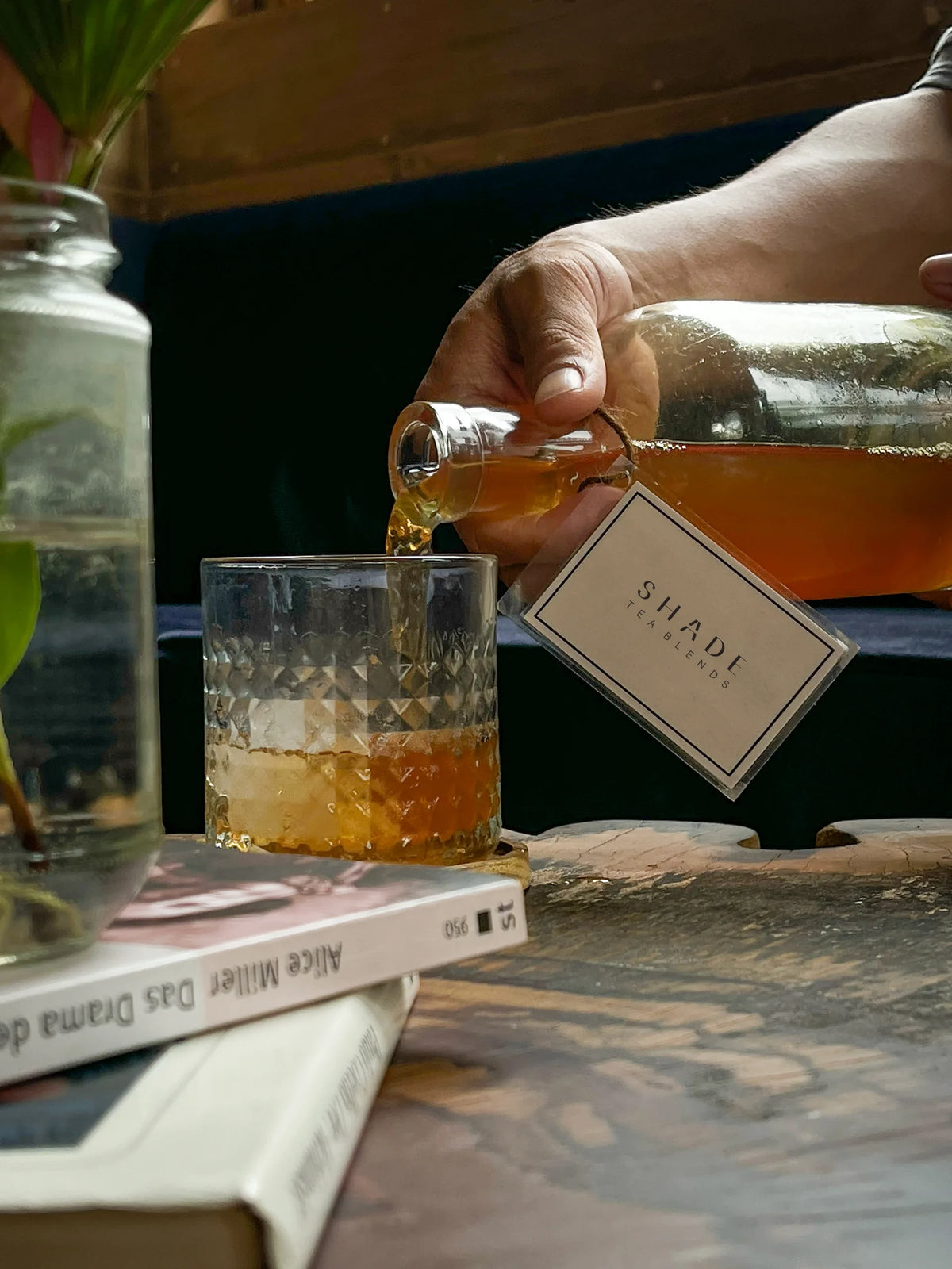 Hand pouring a tea from a bottle labeled SHADE Botanicals Tea Blend into a glass on a rustic surface