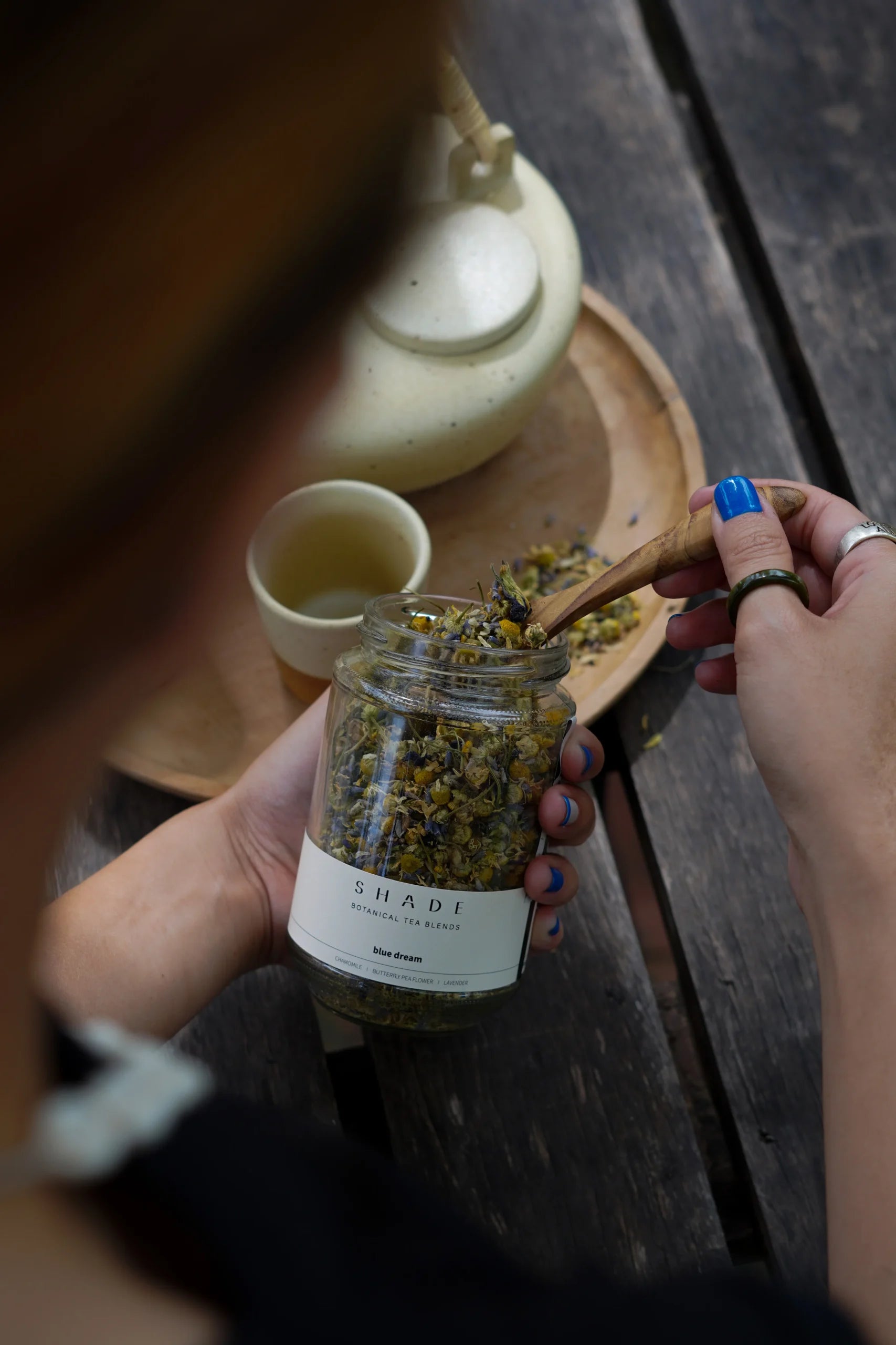 Person holding a jar of Shade Botanicals Tea Blend with a wooden spoon, blurred background