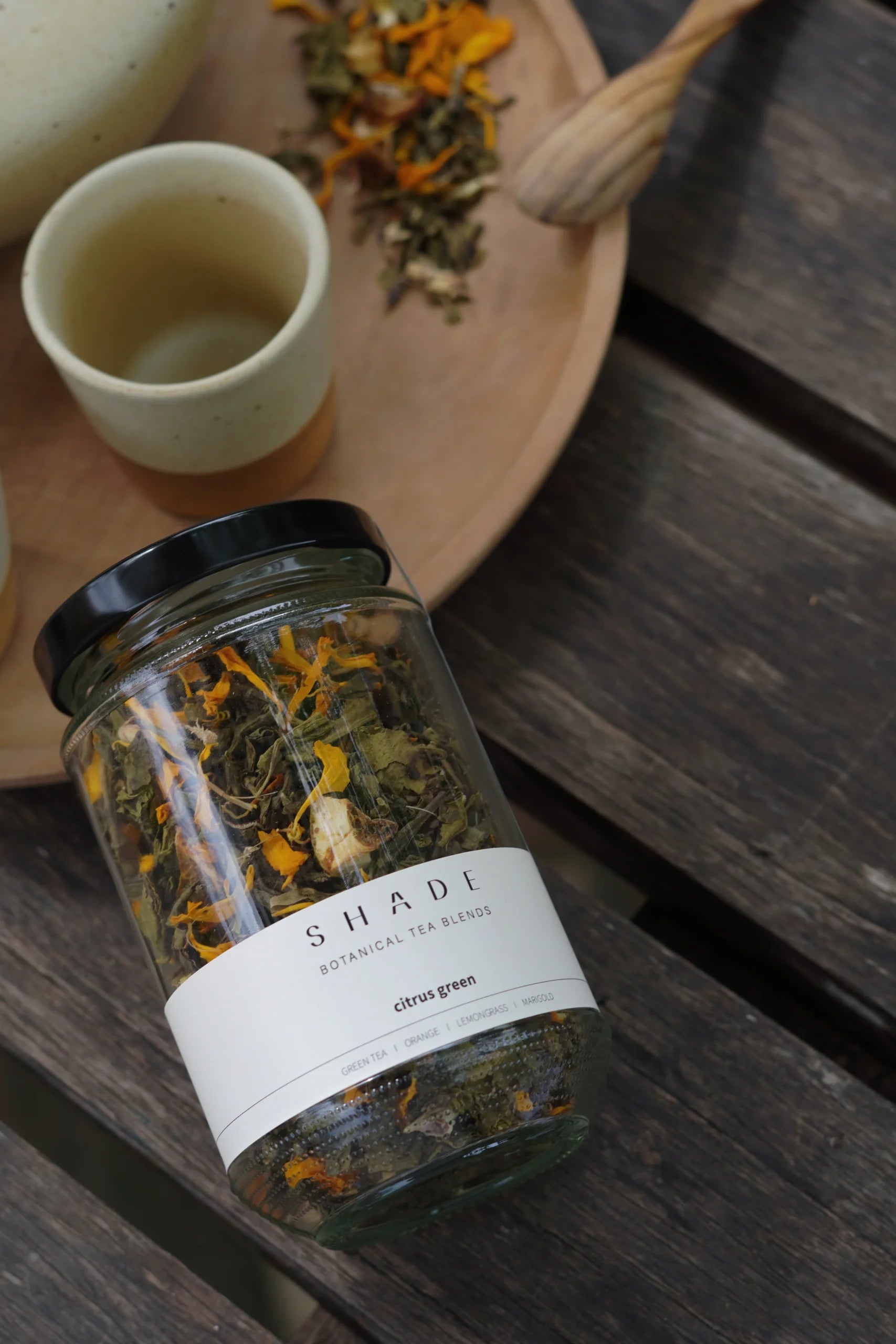 Jar of Shade Botanicals Tea Blend labeled 'Shade' on a wooden surface with a small cup and spoon.