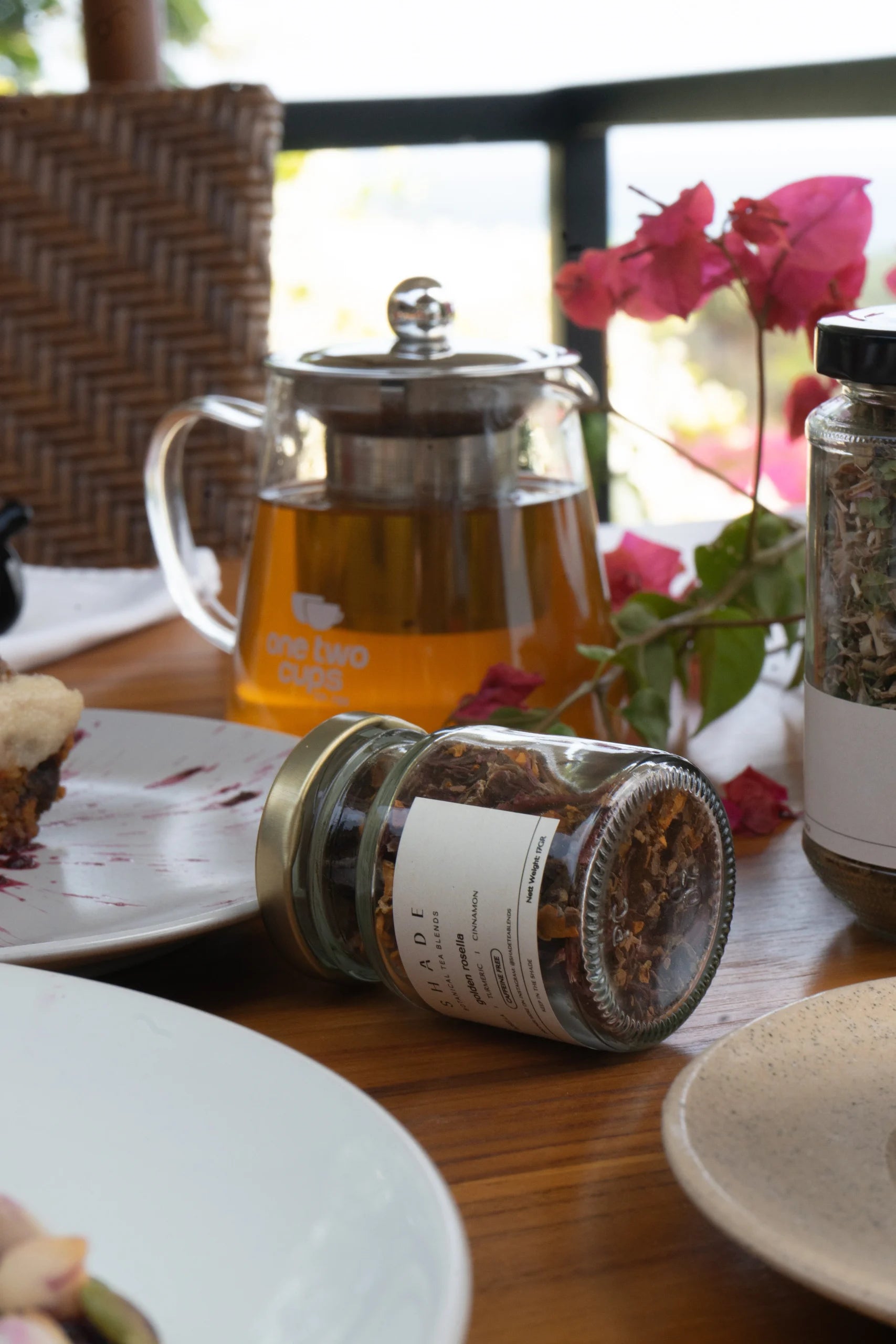 Tea set with teapot, jars of Shade Botanicals Tea Blend, and a plate with a dessert on a wooden table